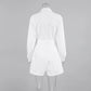 Back view of a white long-sleeved collared playsuit featuring a belted waist and tailored short bottoms, presented on a studio grey background.