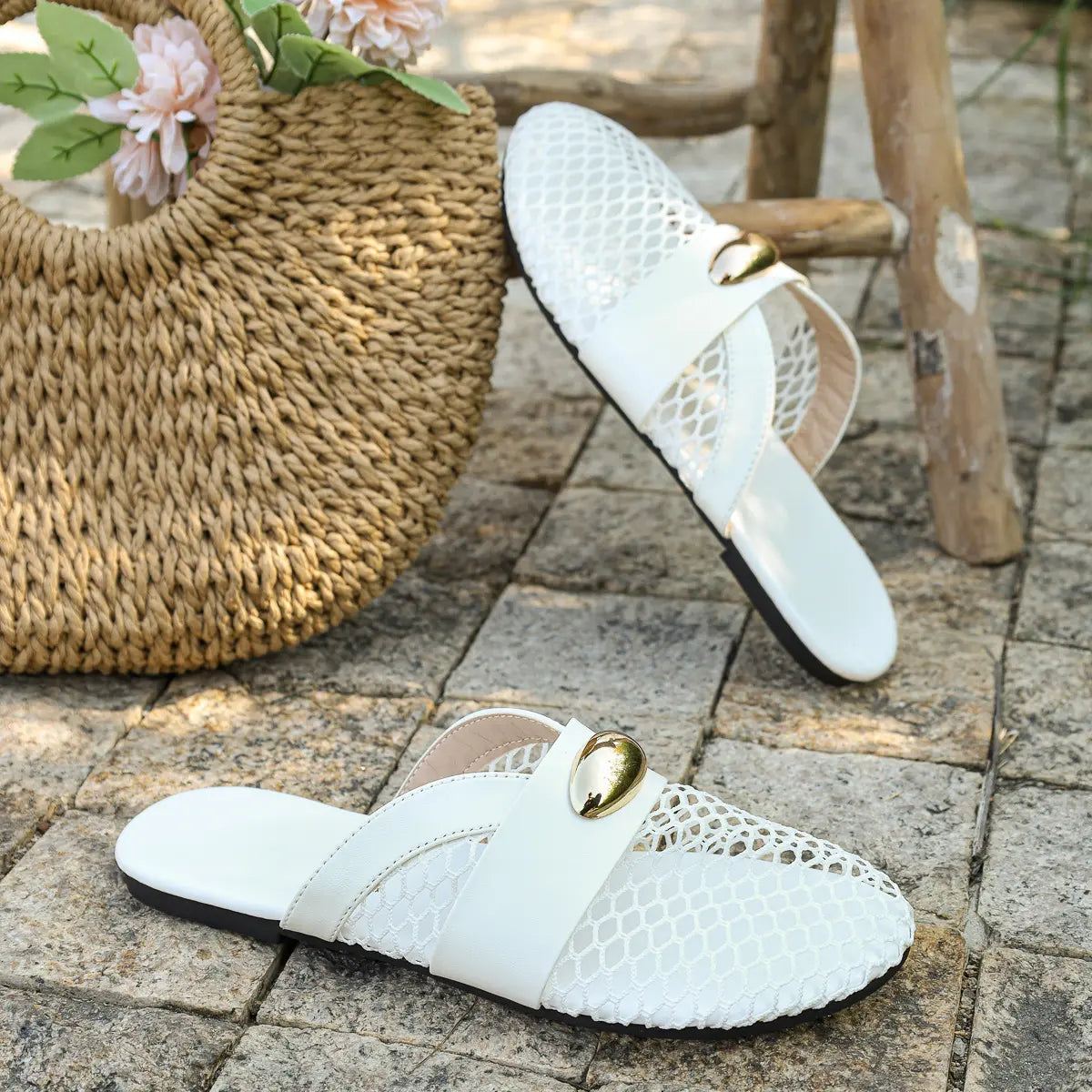 Pair of white mesh pointed-toe flat mules with gold oval hardware and faux leather straps, styled outdoors with a woven straw bag and flowers.