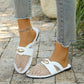A woman's feet wearing white mesh pointed-toe flat mules with a gold oval accent and faux leather strap, paired with distressed jeans, outdoors on a stone path.