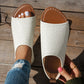 White perforated PU leather flat peep-toe sandals with a tan lining, shown on a woman's foot paired with ripped jeans, with a hand holding the second sandal on a wooden surface.
