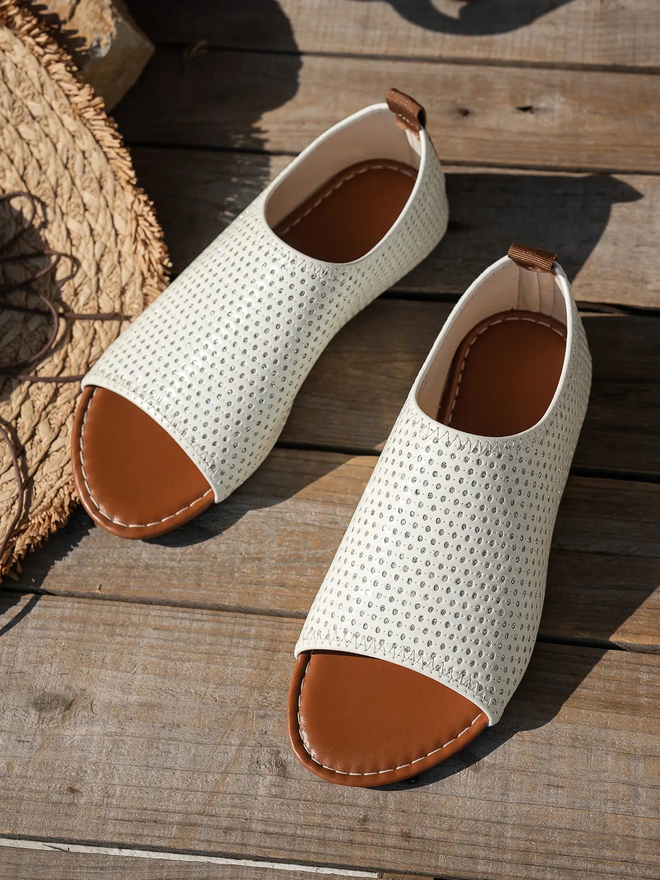 Pair of white perforated PU leather women's flat sandals with a peep-toe and tan lining, styled on a rustic wooden surface next to a straw hat.