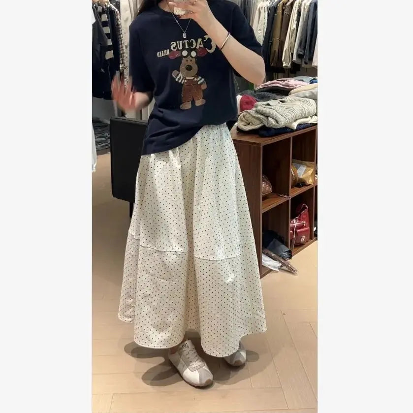 Elegant white A-line midi skirt with classic black polka dots and a distinctive tiered horizontal seam, worn by a model in a sophisticated indoor setting, showcasing its flowing silhouette.