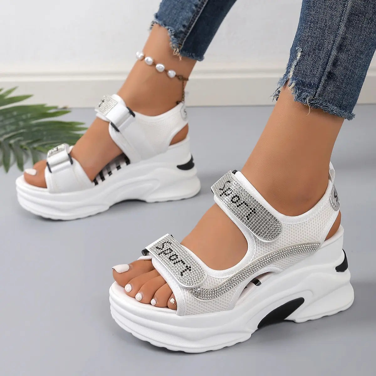 White embellished platform sandals with 'Sport' text on the straps, shown on a woman's feet with ripped jeans and a pearl anklet, offering a sporty chic summer look.