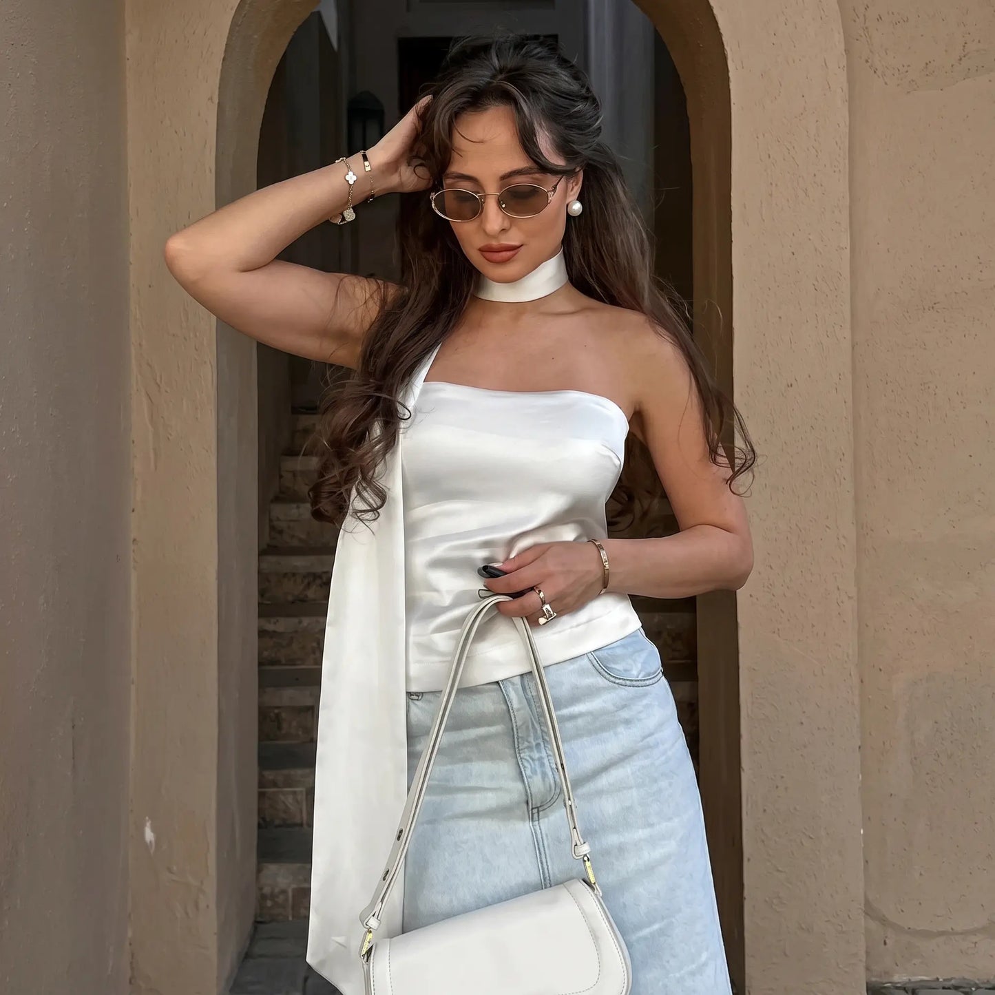 A woman models a white satin bandeau top with an integrated halter scarf, paired with light-wash denim jeans, showcasing elegant summer styling. The background features an arched architectural setting.