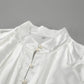 Close-up of a white satin blouse showcasing its high stand collar, cascading ruffled front detailing, and faux pearl button placket. The lustrous fabric and intricate design are highlighted from a detailed angle.