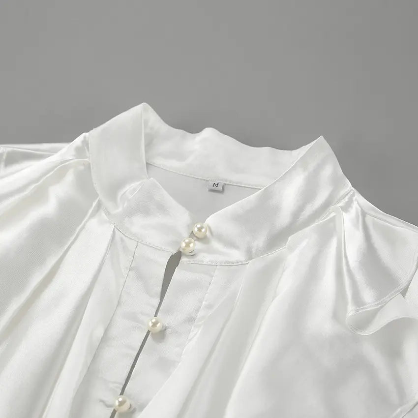 Close-up of a white satin blouse showcasing its high stand collar, cascading ruffled front detailing, and faux pearl button placket. The lustrous fabric and intricate design are highlighted from a detailed angle.