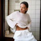 Female model wears a Zomari Pristine White semi-sheer knit sweater with flowing bell sleeves, adjusting its relaxed hemline in an indoor setting. The wide boat neck and dropped shoulders enhance its effortless bohemian style.