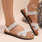 A woman's feet wearing white flat sandals with shimmering silver embellished crossover straps and a delicate pearl anklet, styled against a softly lit background with a sequined accent.
