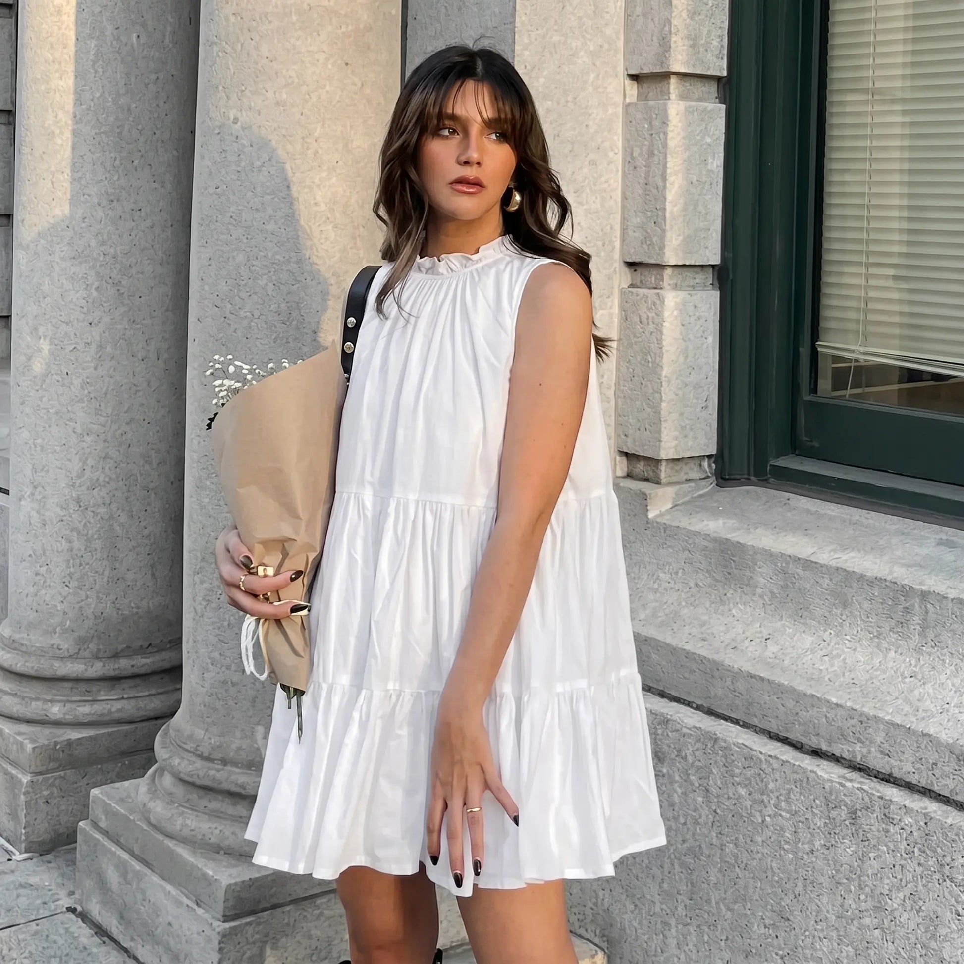 A woman models a white sleeveless tiered mini dress with a ruffled high neckline, posed outdoors against an architectural backdrop, evoking a romantic summer aesthetic. The dress features graceful volume from its tiered construction.