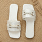 Elegant white slide sandals with square toes and a single wide strap featuring a gold-tone braided buckle and embossed floral pattern, shown on a textured natural mat with a straw hat accent.