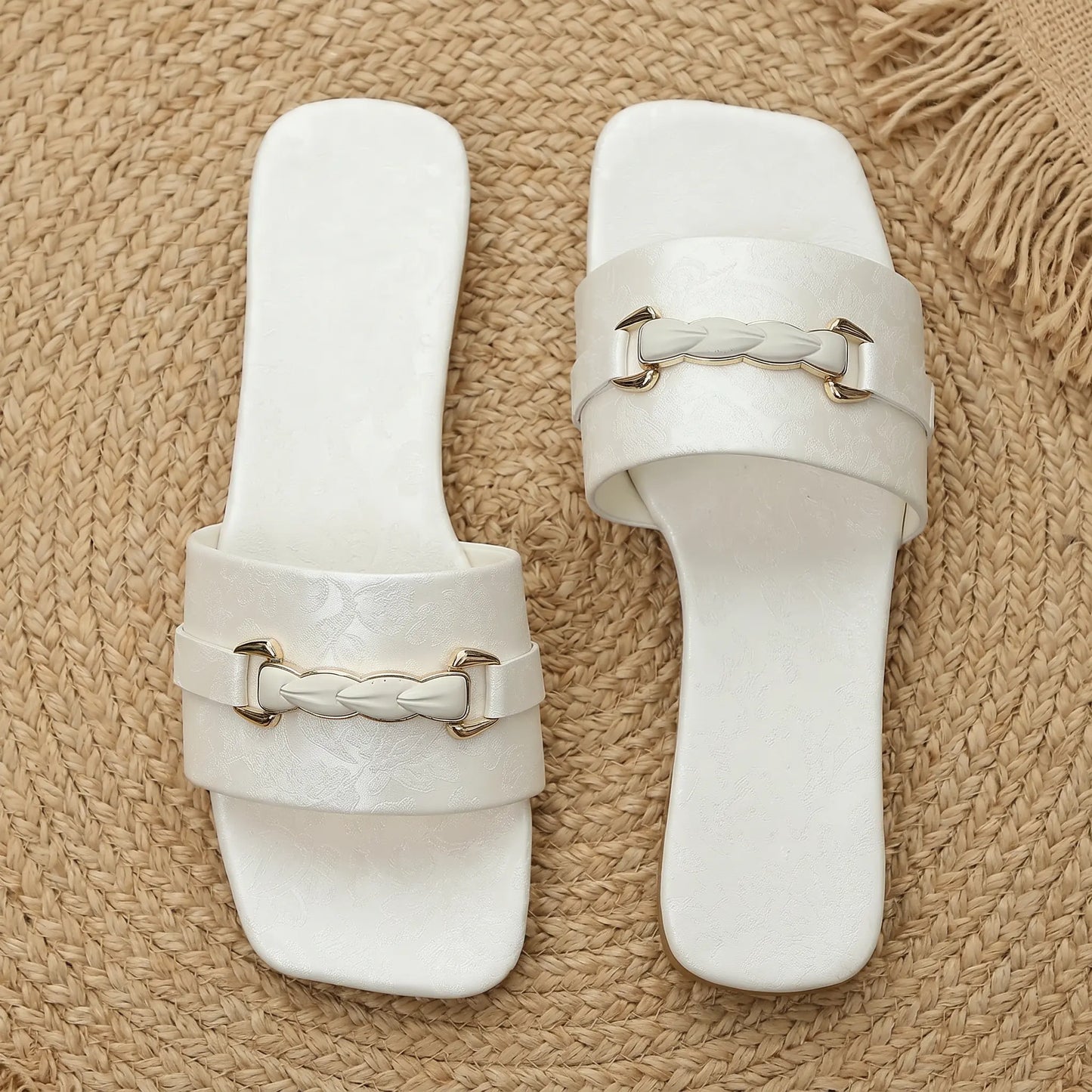 Elegant white slide sandals with square toes and a single wide strap featuring a gold-tone braided buckle and embossed floral pattern, shown on a textured natural mat with a straw hat accent.