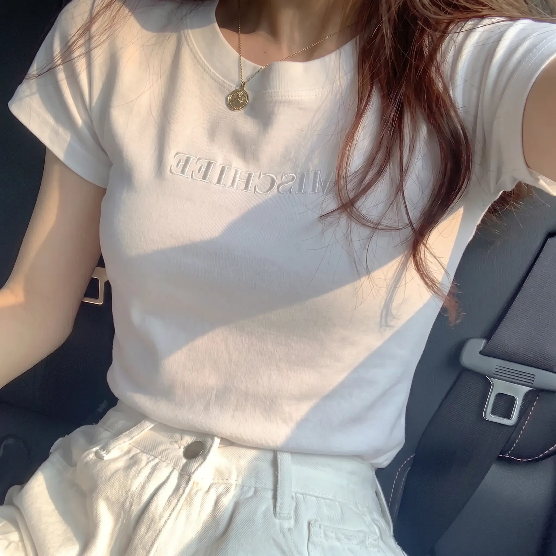 White slim-fit jersey t-shirt with subtle tonal 'MISCHIEF' embroidery, worn by a woman with a gold necklace in a car, highlighting its elegant fit and soft texture.