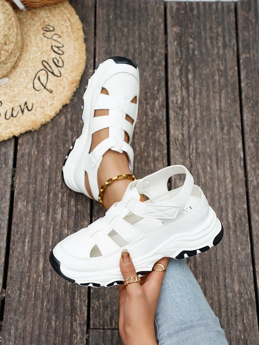 White sporty chic chunky platform sandals with open-back and cut-out straps, styled on feet with denim jeans and a straw hat, set against a wooden background.