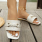 Elegant white square-toe flat slide sandals with a textured strap and gold braided buckle, shown on a model's feet on a wooden deck.