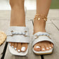 Elegant white square-toe slide sandals featuring a distinctive gold-tone braided buckle and subtle embossed floral pattern, worn on feet with a white pedicure.