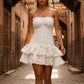 Elegant woman strolling through a sunlit alley wearing a white Taziro strapless smocked mini dress, featuring a tiered broderie anglaise skirt with scalloped edges.