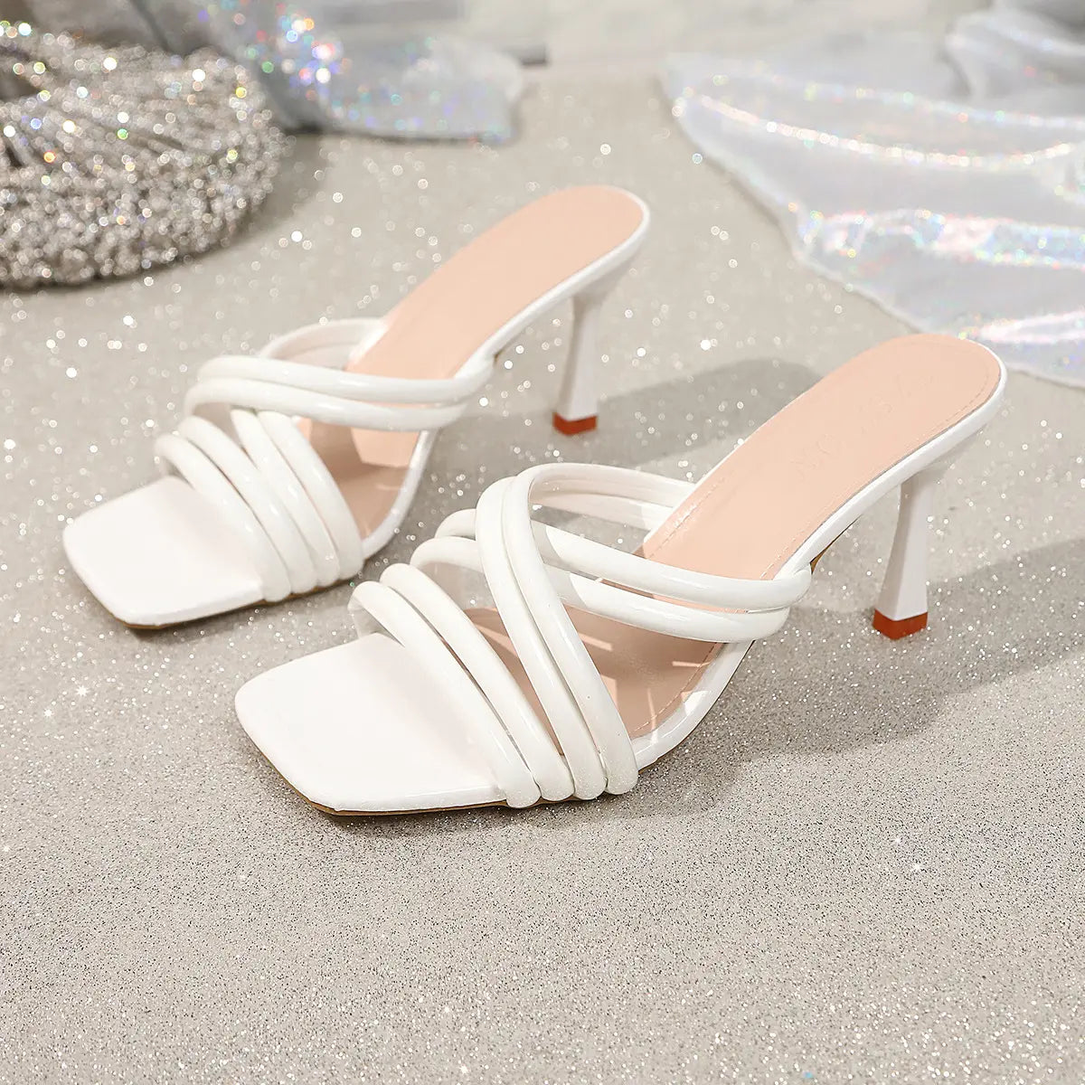White strappy heeled square toe mules featuring glossy PU crisscross straps and a flared sculptural heel, displayed on a sparkly textured surface with draped fabric. Includes embossed 'FASHION' text on the insole.