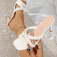 Close-up of a white, high-shine PU leather strappy heeled sandal with a square toe, worn on a foot. Features multiple thin crisscross straps and a flared sculptural heel, shown on a sparkling studio background.