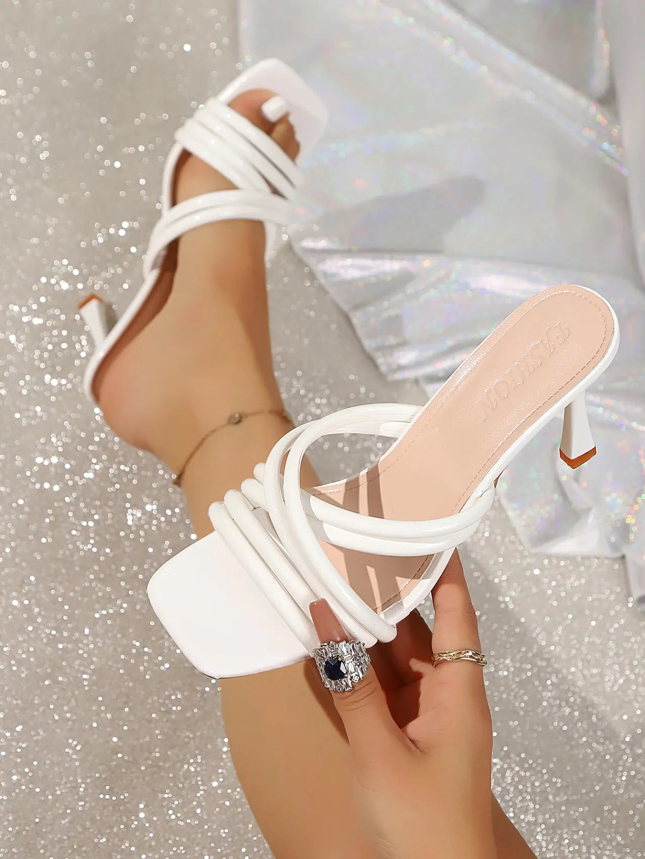 Close-up of a white, high-shine PU leather strappy heeled sandal with a square toe, worn on a foot. Features multiple thin crisscross straps and a flared sculptural heel, shown on a sparkling studio background.