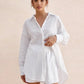 A woman models a white textured cotton shirt and matching shorts set, showcasing its oversized relaxed fit, camp collar, and elasticated waistband, against a soft blush pink background.