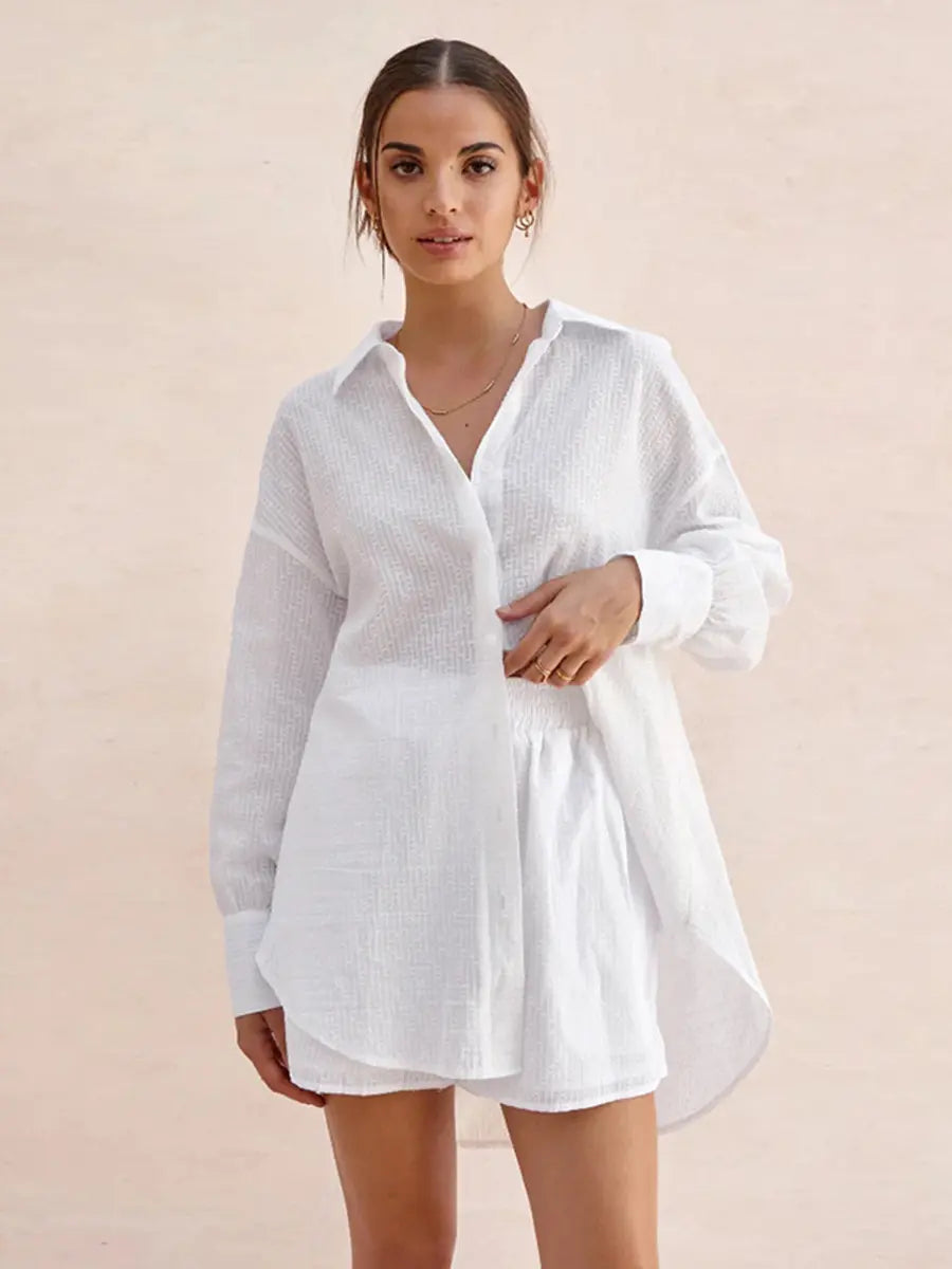 A woman models a white textured cotton shirt and matching shorts set, showcasing its oversized relaxed fit, camp collar, and elasticated waistband, against a soft blush pink background.
