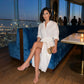 Elegant woman in a white tie-waist midi shirt dress with rolled sleeves and draped tulip skirt, seated at a luxurious rooftop bar.