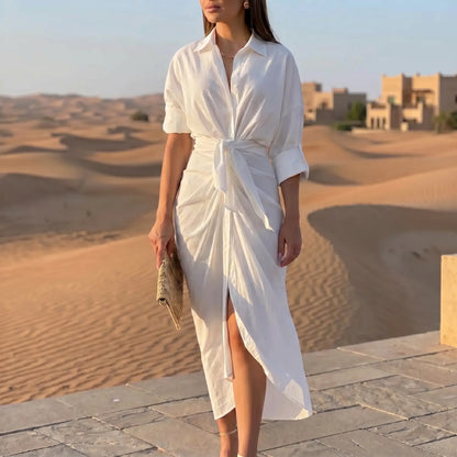 Elegant white tie-waist midi shirt dress with a collared neckline, rolled sleeves, and draped tulip skirt, worn by a model in a sunlit desert scene.
