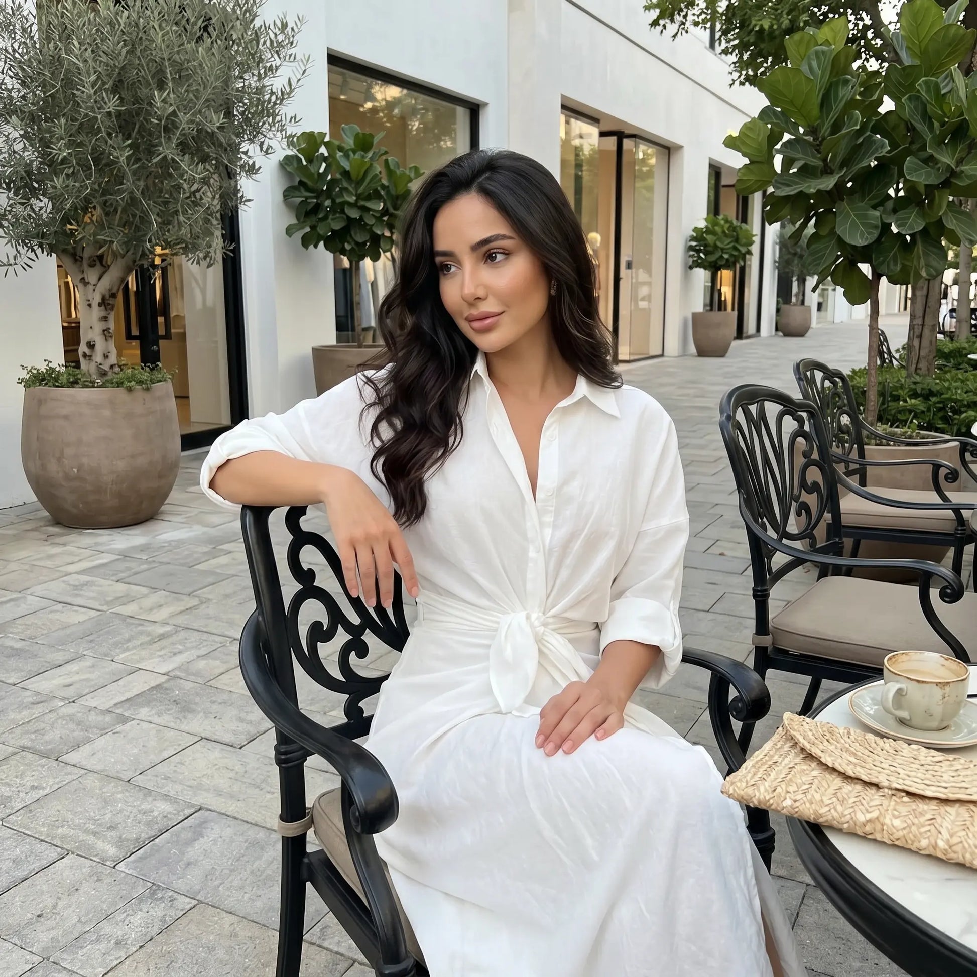 Elegant white tie-waist midi shirt dress with rolled sleeves, worn by a model relaxing at an outdoor cafe. The dress features a pointed collar and a flowing tulip skirt.