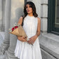 A woman models a white sleeveless tiered mini dress with a ruffled high neckline, holding a bouquet of roses in an outdoor urban setting.