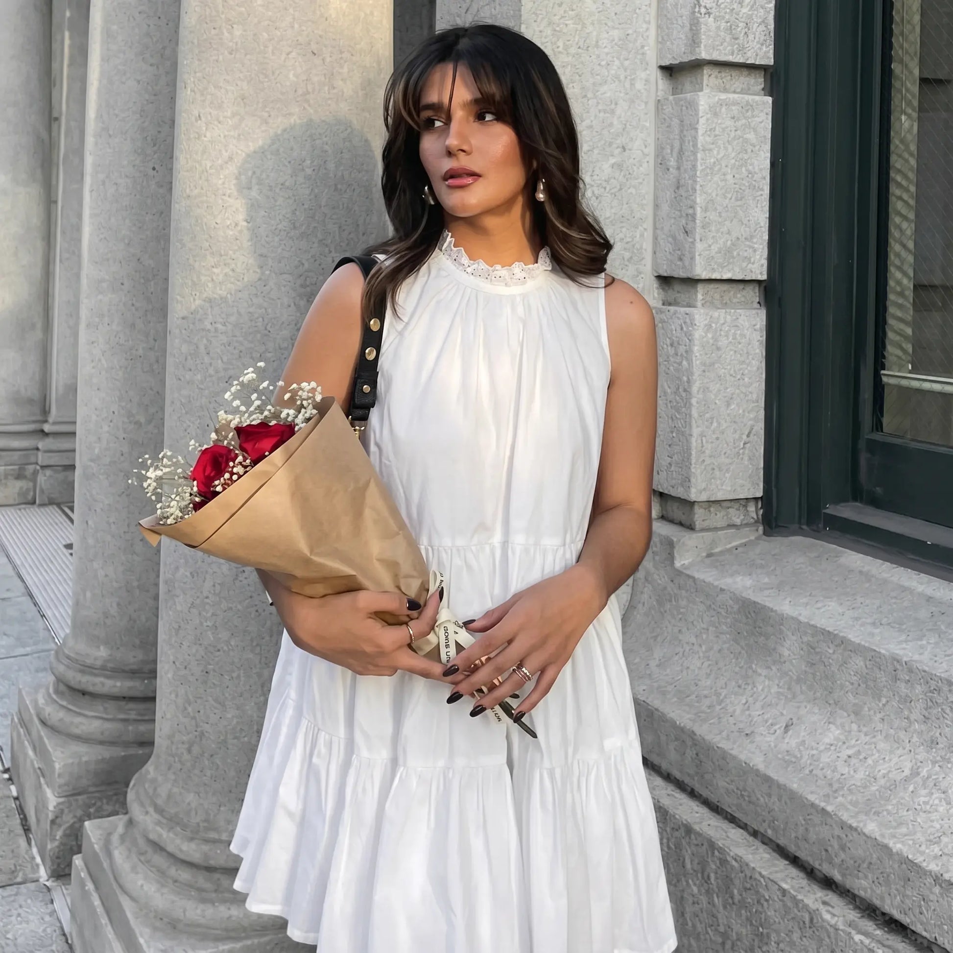 A woman models a white sleeveless tiered mini dress with a ruffled high neckline, holding a bouquet of roses in an outdoor urban setting.
