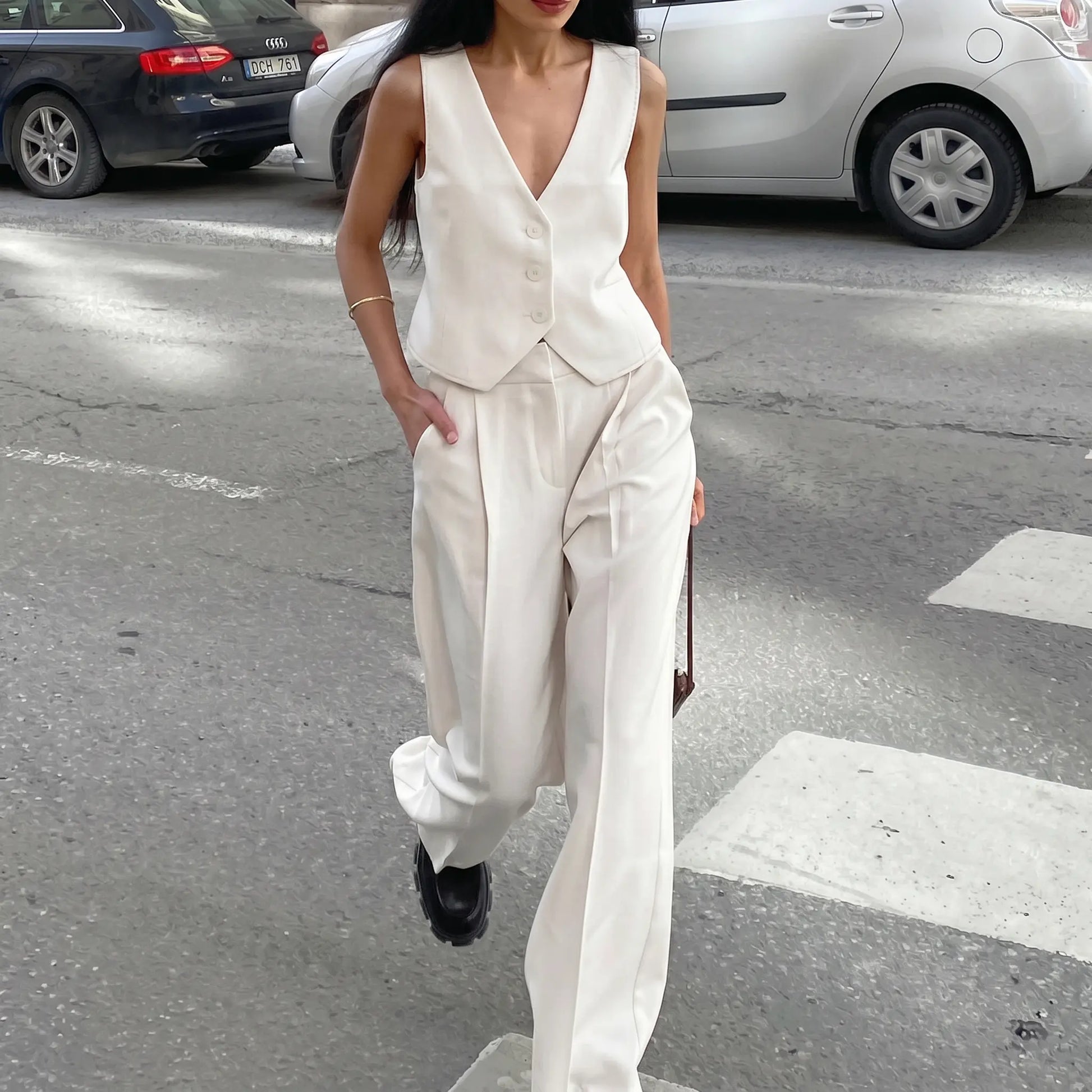 Woman modeling a pristine white sleeveless V-neck vest and fluid wide-leg trousers ensemble, walking across a city street with a subtle sheen to the fabric.