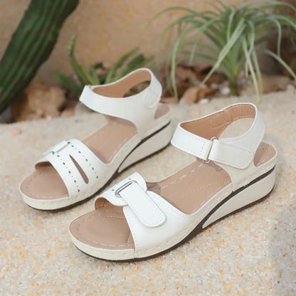 White Lyraia women's low wedge sandals with peep-toe, perforated front straps, and adjustable hook-and-loop closures, set against a sandy background.