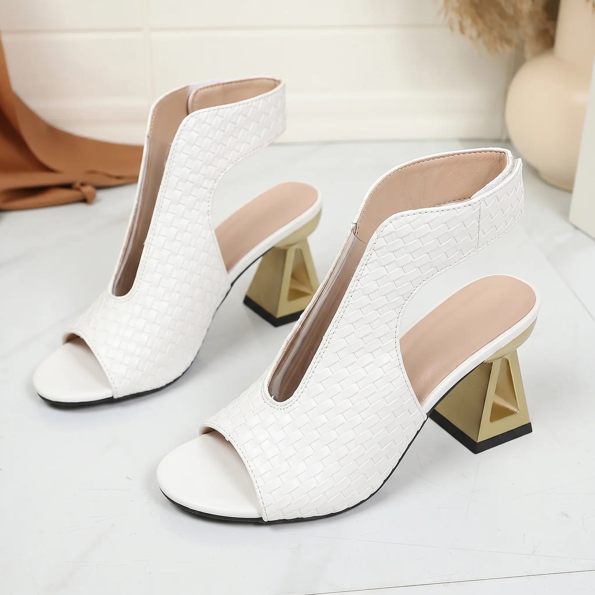 White Aethel peep-toe block heel sandals featuring a woven texture upper and a distinctive geometric gold heel, with a delicate chain ankle strap.