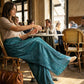 Woman wearing deep teal wide leg pleated plissé trousers with a high-waist fit, sitting at a cafe table.