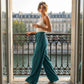 Woman wearing deep teal wide leg pleated trousers with a high-waist fit, standing on a balcony with a city view.