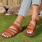 Brown women's flat braided strap slide sandals worn by a model with white-toe nails and a turquoise anklet, set in a natural outdoor scene with grass and rocks.