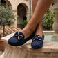 Women's navy blue faux-suede moccasin loafers with gold metallic bows, featuring raised toe stitching, worn by a person with crossed legs on a marble fountain edge in a sunny European courtyard setting.