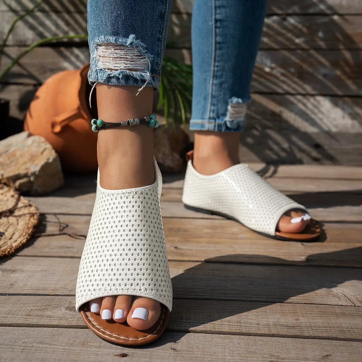 Women's white perforated PU leather flat peep-toe sandals worn with distressed denim jeans, shown in a casual lifestyle setting on a wooden deck. A beaded anklet adorns one foot.