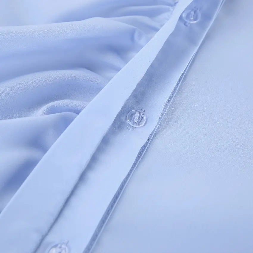Close-up view of a powder blue collared blouse, showcasing the asymmetrical ruffle detail along the front placket and clear buttons. The smooth, subtle sheen fabric highlights the quality craftsmanship.