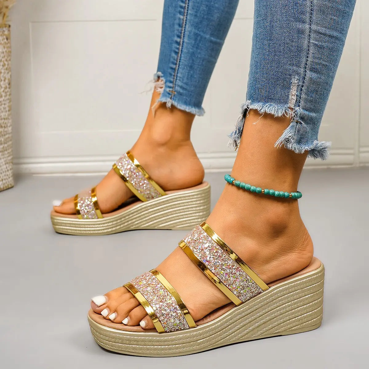 Women's gold glitter wedge slide sandals with two wide, sparkling straps and a textured wedge heel, worn by a model with distressed denim jeans.