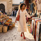 A woman strolls through a vibrant outdoor market, showcasing a flowing cream midi dress and eye-catching red stiletto sandals with a cross-strap detail. She carries a woven orange tote bag and wears large gold hoop earrings. She is browsing market stalls filled with textiles and baskets, embodying a bohemian summer style.