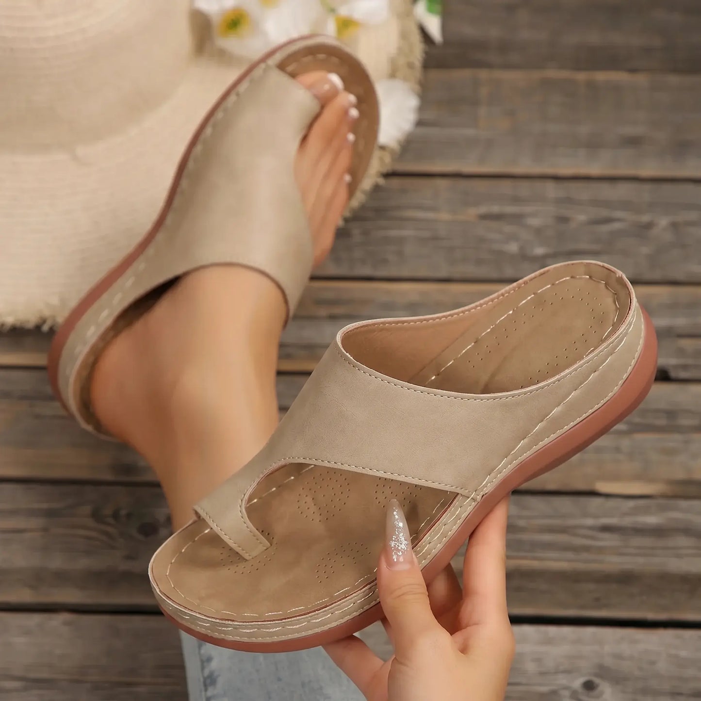 A pair of women's casual slip-on slides in sandstone beige, featuring a PU leather upper, toe-post design, and a contoured footbed on a rustic wooden surface with a straw hat.
