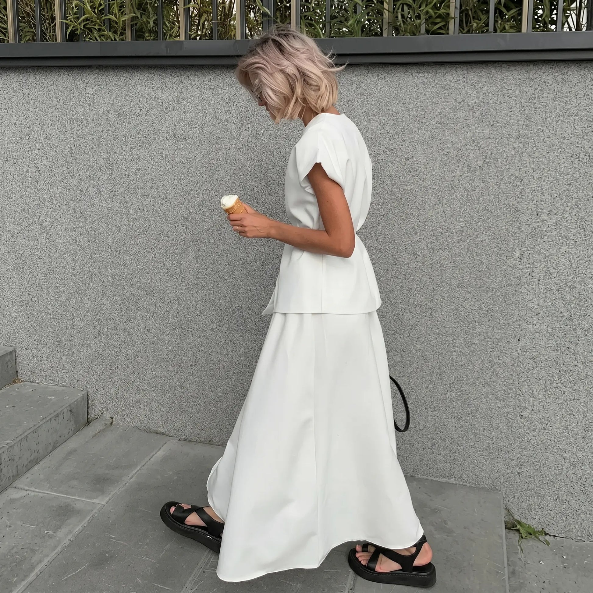 Elegant white V-neck sleeveless top paired with a flowing A-line midi skirt, worn by a woman walking outdoors with an ice cream cone. The sophisticated two-piece set showcases a timeless design with a matte finish.