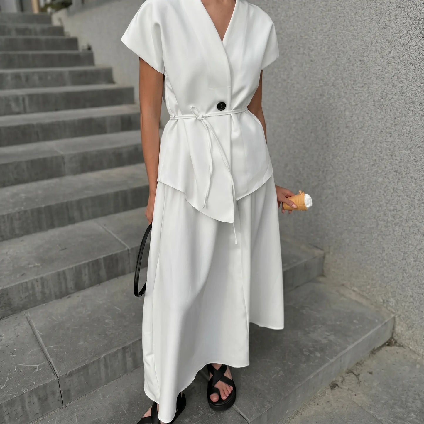 A woman models an elegant white sleeveless V-neck two-piece set featuring a V-neckline, asymmetrical overlay top with a tie belt, and a flowing A-line midi skirt, styled with black sandals on concrete stairs.