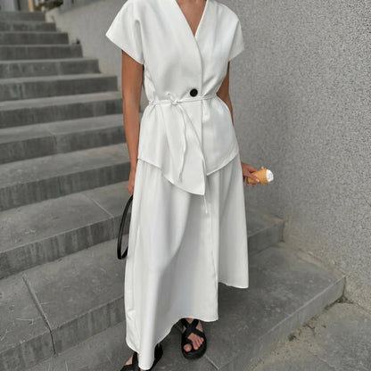 A woman models an elegant white sleeveless V-neck two-piece set featuring a V-neckline, asymmetrical overlay top with a tie belt, and a flowing A-line midi skirt, styled with black sandals on concrete stairs.