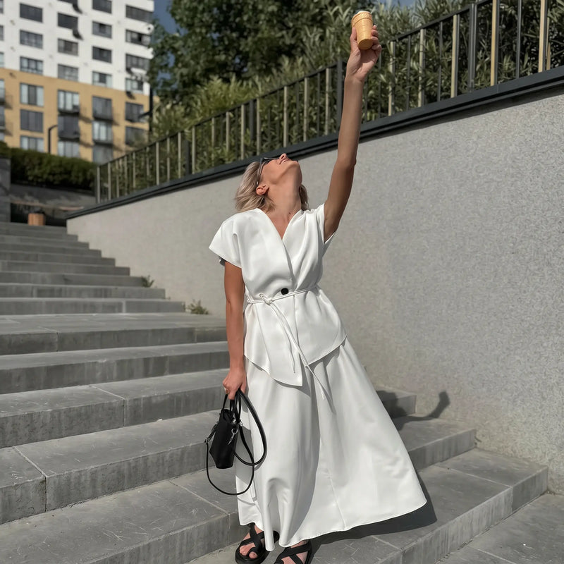 Elegant white two-piece set on a woman posing on outdoor steps, featuring a V-neck sleeveless top with asymmetrical overlay and tie belt, paired with a flowing A-line midi skirt. She holds a black handbag and wears black sandals.