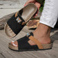 Xylia embellished flat slide sandals in Onyx Black, featuring a natural cork footbed, tan accent strap, and clear crystal butterfly embellishment, shown on a foot with distressed white jeans.