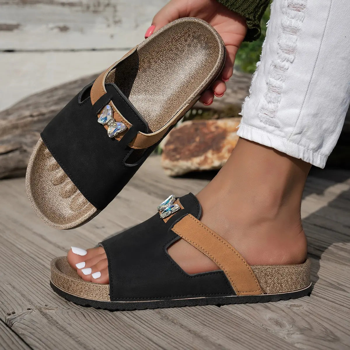 Xylia embellished flat slide sandals in Onyx Black, featuring a natural cork footbed, tan accent strap, and clear crystal butterfly embellishment, shown on a foot with distressed white jeans.