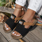 Xylia black embellished slide sandals with tan accent strap and crystal butterfly detail, worn on feet with white jeans and a seashell anklet on a wooden deck.