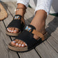 Close-up of Xylia embellished flat slide sandals worn by a person with white nail polish, featuring a black suede-effect strap with a crystal butterfly and a tan accent strap on a wooden deck.
